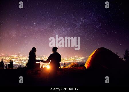 Happy couple in silhouette sitting near campfire and orange tent. Night sky with milky way stars and city lights at background. Stock Photo