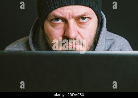 Man wearing black cap and hoodie sitting behind the computer monitor in the dark, close up, hacking computer system concept Stock Photo