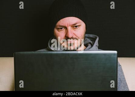 Man wearing black cap and hoodie sitting behind the computer monitor in the dark, close up, hacking computer system concept Stock Photo