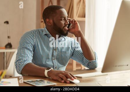 Happy afro american businessman in shirt talking by phone with client and smiling while working on the computer at his working place at home. Freelance. Home office. Working remotely Stock Photo