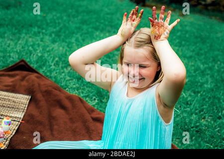 Cute little girls shows her dirty colored with paints palms sitting on blanket on grass in backyard. Stock Photo