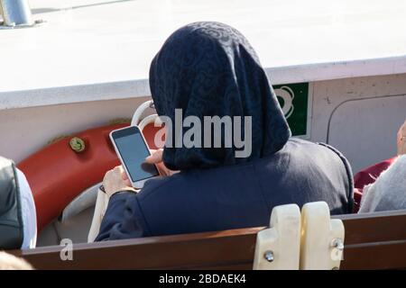 Woman in headscarf is dealing with mobile phone. The photo was taken on the ferry and sunny outdoors. Stock Photo