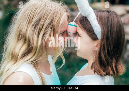 Cute little sisters wearing bunny ears on Easter day d playing with painted egg holding it between thier foreheads. Close up shot. Stock Photo