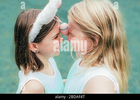 Cute little sisters wearing bunny ears on Easter day d playing with painted egg holding it between thier foreheads. Close up shot. Stock Photo