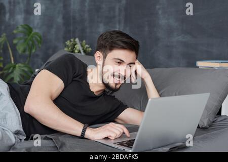 Smiling handsome young man lying in bed and using laptop while hatting with friends vie online app Stock Photo