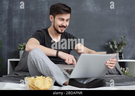 Positive young bearded man sitting with crossed legs on bed and eating chips while talking to relatives via video link on laptop at quarantine Stock Photo