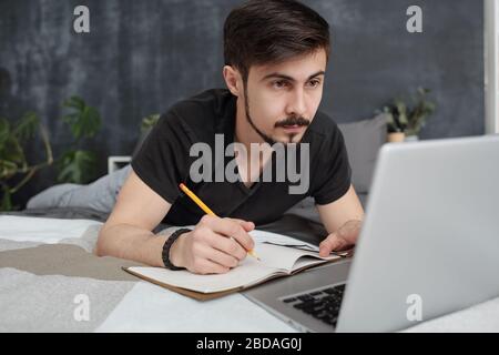 Concentrated young bearded student lying in bed and making notes while listening to lecture online at quarantine Stock Photo