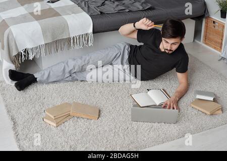 Pensive young student in homewear lying among books on carpet analyzing online information on laptop while doing coursework Stock Photo