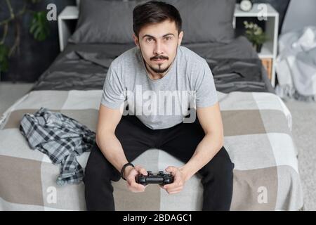 Curious young man sitting on bed and using joystick while playing video game at home during quarantine Stock Photo