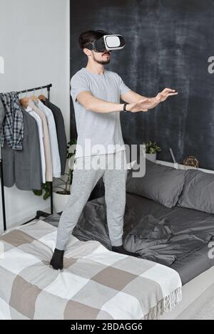 Young bearded man in homewear standing on bed and gesturing hands while playing video game in virtual reality goggles Stock Photo