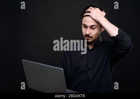 Puzzled young bearded programmer in black shirt holding hand on head while looking at information on laptop screen Stock Photo
