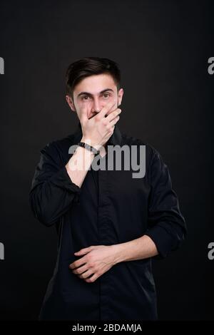 Portrait of shocked young brunette man in black shirt covering mouth with hand, not believing his eyes Stock Photo