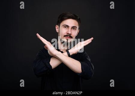 Portrait of young bearded man standing against black background and crossing arms while making stop gesture Stock Photo