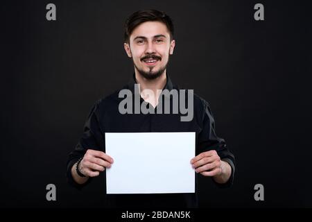 Portrait of smiling handsome young bearded man in black shirt holding blank paper in hands against dark background Stock Photo