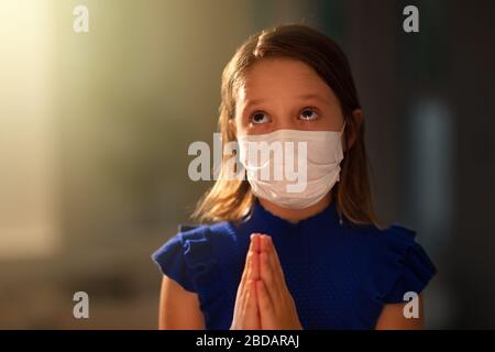 Pray for the sick. Child in face mask praying. Little girl in hospital chapel or church during coronavirus outbreak. Virus pandemic. Kids worship. Chi Stock Photo