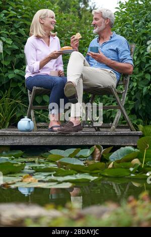 Mature Couple Enjoying Afternoon Tea And Cake Sitting On Chairs On Wooden Jetty By Lake Stock Photo