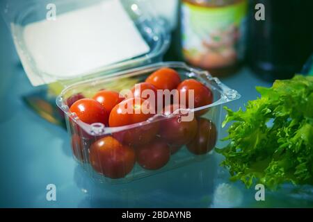 In the refrigerator, on a glass shelf, there is a plastic bag of ripe cherry tomatoes, and lettuce leaves are next to it. In the background are jars o Stock Photo