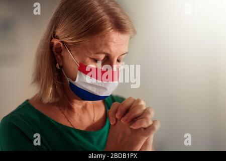 Pray for the Netherlands. Woman in face mask praying for Holland. Senior Dutch patient in hospital chapel or church during coronavirus outbreak. Virus Stock Photo