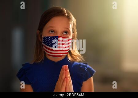 Pray for America. Child in face mask praying for USA. Little American girl in hospital chapel or church during coronavirus outbreak. Virus pandemic. K Stock Photo