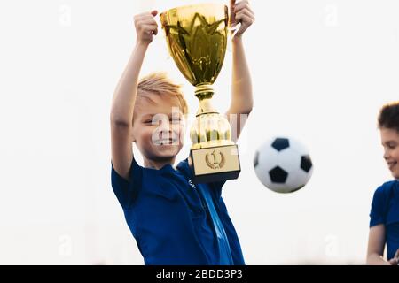 Happy boy rising golden trophy. Child winning sports competition. Overjoyed kid holding golden prize award. Soccer ball in the background Stock Photo