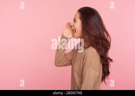 Attention, important message! Side view of young woman with brunette wavy hair shouting news or proclaiming idea with hand near mouth, announcing adve Stock Photo