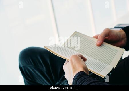 closeup of a young caucasian man, in casual wear, reading a book sittin in an outdoors armchair on the balcony Stock Photo