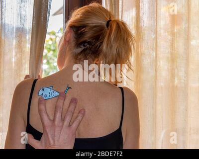 A man sticks an April fool's day fish of colored paper on the back of a woman looking out the window Stock Photo