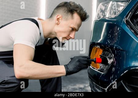 Car detailing wash. Detailing cleaning with soap at car wash. Young man in rubber gloves washes car radiator grille of luxury blue car with special br Stock Photo