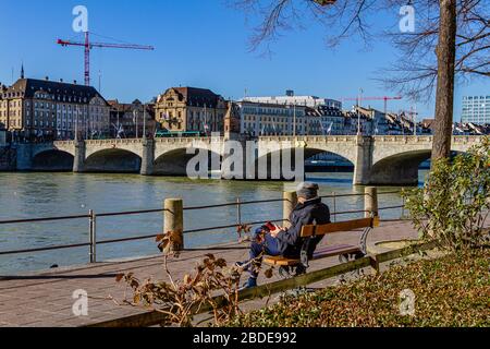 A person sitting on a bench beside the river Rhine and the historic Middle Bridge in the city of Basel, Switzerland. February 2020. Stock Photo