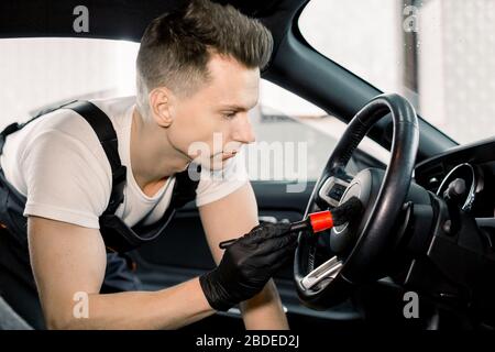 Side view close up portrait of young Caucasian male car wash detailing worker using cleaning brush and removing dust from car steering wheel. Car deta Stock Photo