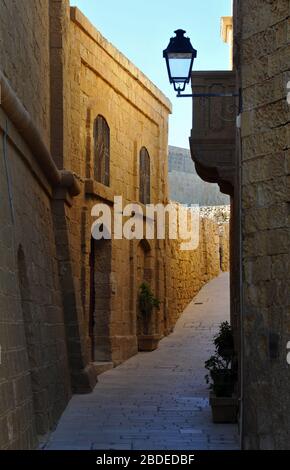 Stone buildings on a quiet path lit by the setting sun in the historic Citadel of Victoria (Rabat), the capital city of the island of Gozo in Malta.