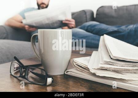 man relaxing on sofa reading a newspaper with stack of newspapers and coffee mug in foreground Stock Photo