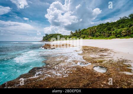 Rocky old reef coastline on tropical Police Bay beach with rolling waves and white clouds on Mahe Island, Seychelles Stock Photo