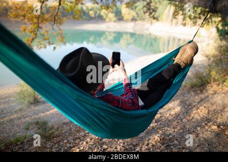 Italy, Man lying in hammock near lake and using smart phone Stock Photo