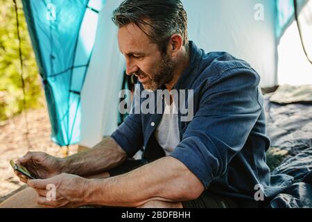 Mature man sitting in a tent and looking at his mobile phone. Mid adult camper using smart phone at campsite. Stock Photo