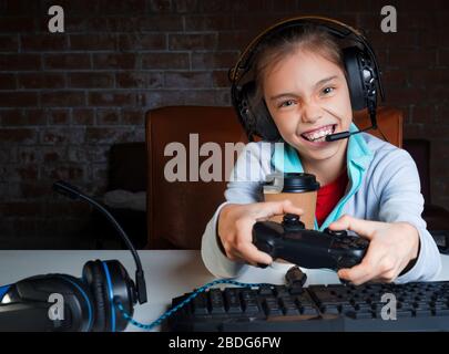 A young girl in a big headphones with microphone is sitting in front of a monitor and playing video games with a happy excited face. Win. Stock Photo