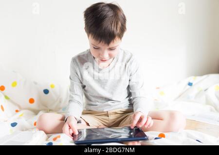 Game. Boy plays with smartphone. School Boy on bed at home with digital pad tablet in hand, doing homework. Distance learning online education Stock Photo