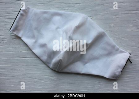White handmade fabric cotton face mask with black elastic straps on white wooden background. Reusable protective face mask to help prevent the spread Stock Photo