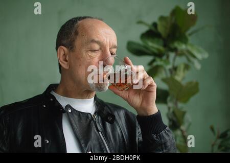 Senior man in black leather jacket is drinking whisky from glass in loft style room with light green walls and houseplants on background. Alcohol Stock Photo