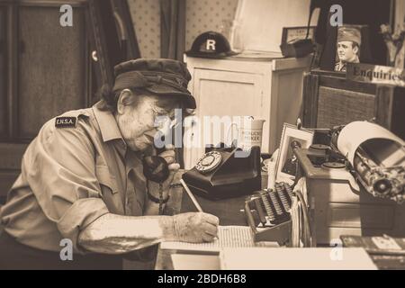 Old-fashioned sepia side view of senior woman in uniform working isolated in vintage office, Severn Valley Railway, 1940s wartime summer event, UK. Stock Photo