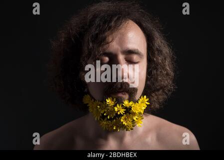 Studio dramatic portrait of a young guy of thirty years old, with his eyes closed. yellow flowers are woven into curly hair and a long beard. Concept Stock Photo