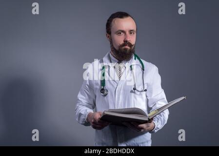 Portrait of a bearded doctor in a white coat and a phonendoscope holding educational books in his hands. Studio photo on a gray background. Self-educa Stock Photo