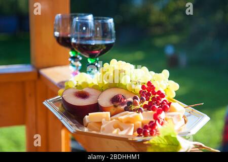 Two glasses of red wine and cheese board, fresh grapes, peaches and berries on wooden table. Outside location Stock Photo