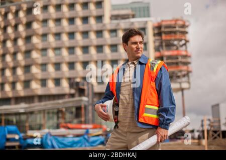 Handsome young architect in a high visibility vest holds his hardhat and building plans on a construction site. Stock Photo