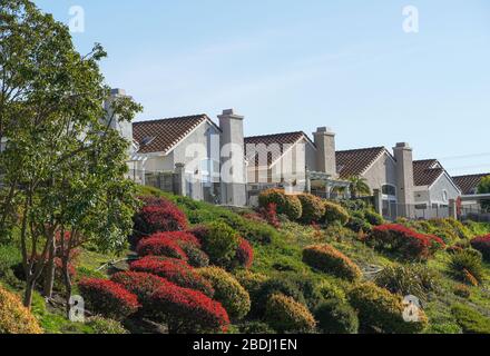 Row of single story stucco houses with red tile roofs and chimneys in a retirement community: light blue sky, copy space, bushes, sloping landscaping. Stock Photo