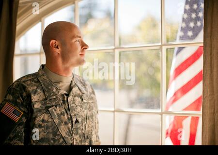 Thoughtful soldier looking through a window while standing inside his home. Stock Photo