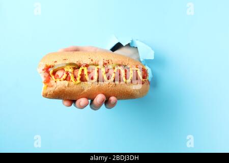 Hand holds hot dog from hole on blue background Stock Photo