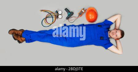 A male builder was left without work in an economic crisis. Unemployment is not work. Male worker lies on his back, near a work tool on a gray background. Stock Photo