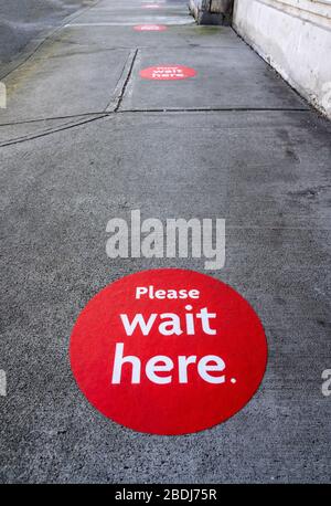 Round red physical distancing notification with words "Please Wait Here" on sidewalk during Coronavirus Covid-19 outbreak Stock Photo