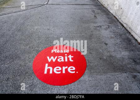 Round red physical distancing notification with words "Please Wait Here" on sidewalk during Coronavirus Covid-19 outbreak Stock Photo